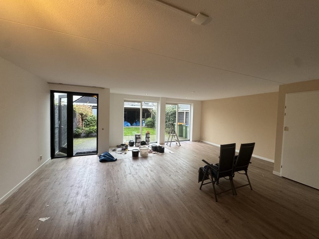 Ruime woonkamer in een bestaande woning met lichte wanden, een houten vloer en tuindeuren, tijdens de werkzaamheden